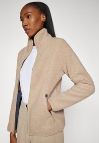 Giacca in pile beige con colletto alto, chiusura a zip frontale e tasche laterali. Tessuto morbido e testurizzato con dettagli a contrasto.
