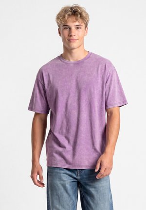 Giovane uomo con capelli biondi corti che indossa una maglietta viola larga e jeans blu, in piedi davanti a uno sfondo chiaro e uniforme.