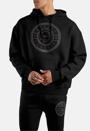 Hombre con sudadera con capucha negra Reebok Classic y pantalones deportivos negros a juego con logos circulares, manos entrelazadas, mirando hacia un lado.