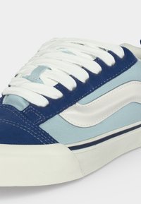 Gros plan d'une basket bleue et bleu clair avec des lacets blancs, une bande latérale blanche et une semelle en caoutchouc blanche avec une ligne bleu marine.