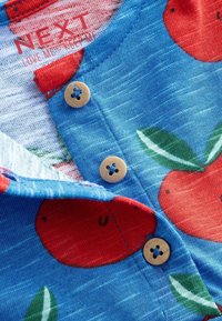 Tissu bleu avec un motif d'oranges rouges et de feuilles vertes, comportant trois boutons beige au niveau de l'encolure. Texture douce et couleurs vives.