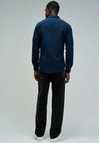 Camisa de botones azul marino con un acabado texturizado, combinada con jeans negros. El modelo lleva zapatillas blancas. Ambos artículos tienen un estilo casual.