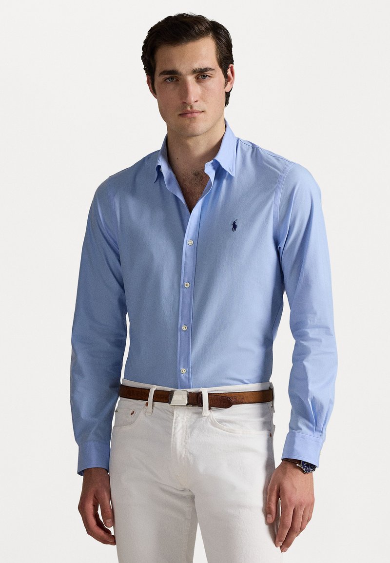 Vīrietis, valkājot gaiši zilu pogā tirkotu kreklu, kas ievilktas baltās biksēs ar brūnu jostu, stāv uz vienkārša baltā fona.