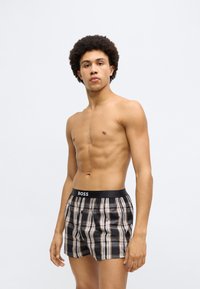 Fekete és bézs kockás boxeralsó fekete elasztikus derékpánttal, amelyen egy 'BOSS' logó látható; egy sportos testalkatú férfi modell viseli.