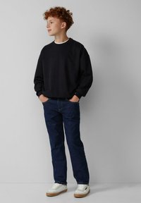 Adolescente con capelli rossi ricci, che sta in piedi con le mani nelle tasche, indossando una felpa nera, jeans blu e sneakers bianche di fronte a un muro semplice.
