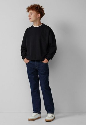 Un adolescent aux cheveux bouclés roux se tient avec les mains dans les poches, portant un sweat-shirt noir, un jean bleu et des baskets blanches contre un mur uni.