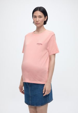 Rase naine, lühikeste mustade juustega, kandes roosat T-särki, millel on kiri "Berry Good Time", ja sinist teksaseelikut, seisab valge tausta ees.