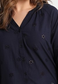 Chemise bleu marine à boutons avec broderie florale, dotée d'une texture douce et d'un col en V. Épaules légèrement froncées, couture visible.