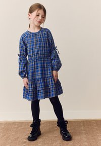 Abito a quadretti blu con maniche a sbuffo, orlo arricciato e dettagli con fiocchi. Abbinato a stivali in pelle nera. Sullo sfondo neutro.