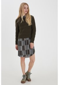 Olivgrüner strukturierter Pullover über einem gemusterten schwarz-weißen Kleid mit Streifen, kombiniert mit olivgrünen Stiefeletten.
