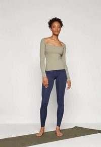 Naine seisab paljajalu rohelisel jooga matil, seljas õhuke oliivivärvi pikkade varrukatega topp ja tumesinised retuusid, tavalise tausta ees.