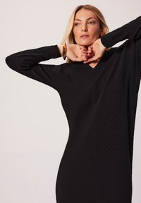 Czarna, prążkowana sukienka z długimi rękawami i dekoltem w kształcie litery V. Tkanina ma strukturalny wzór i dopasowuje się do ciała, podkreślając sylwetkę.