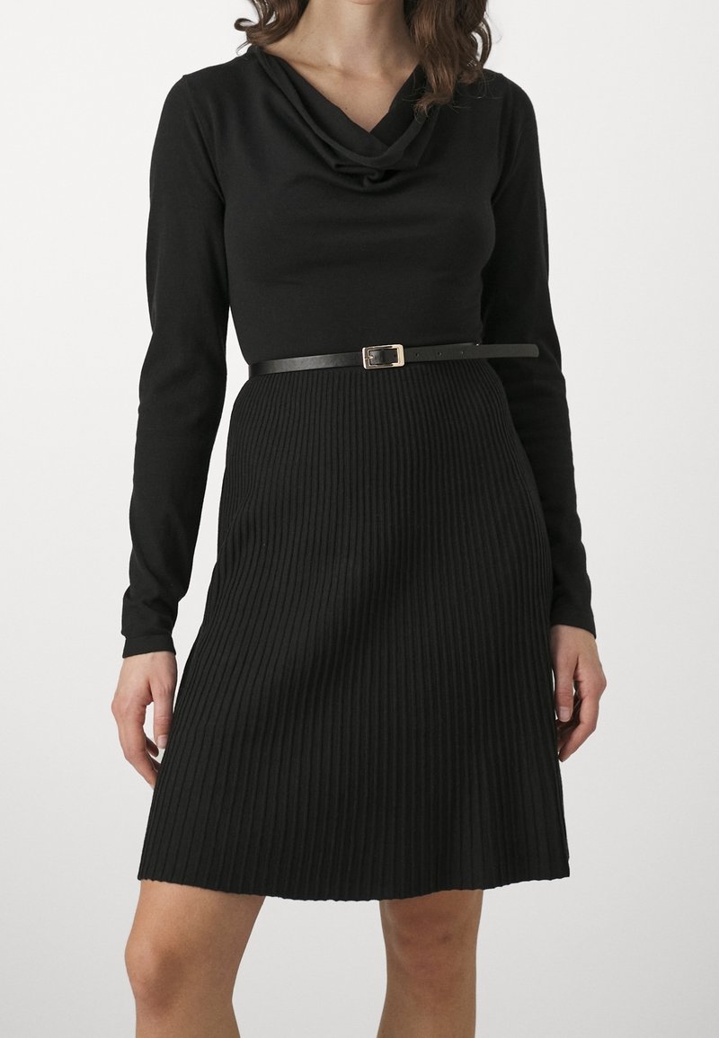 Robe noire à manches longues avec un col bénitier, une texture côtelée et une taille cintrée mise en valeur par une fine ceinture noire avec une boucle dorée.