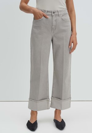 Vrouw draagt lichtgrijze jeans met wijde pijpen en grote omslagen, een wit mouwloos topje ingestopt en zwarte puntige platte schoenen, staand met één hand in haar zak.