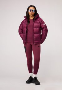 Bordowa kurtka puchowa z kapturem, zestawiona z dopasowaną bluzką i legginsami z czarną boczną kieszenią. Czarne buty sportowe i okulary przeciwsłoneczne.