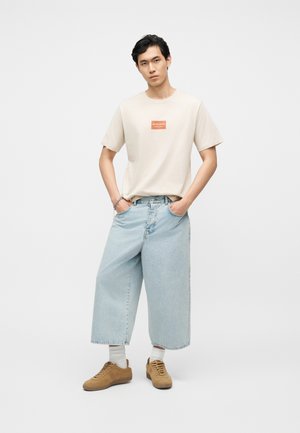 Nuori mies seisoo käsineen taskuissa, päällään beige paita, jossa on logo, leveät vaaleansiniset lyhyet farkut, valkoiset sukat ja ruskeat tennarit.