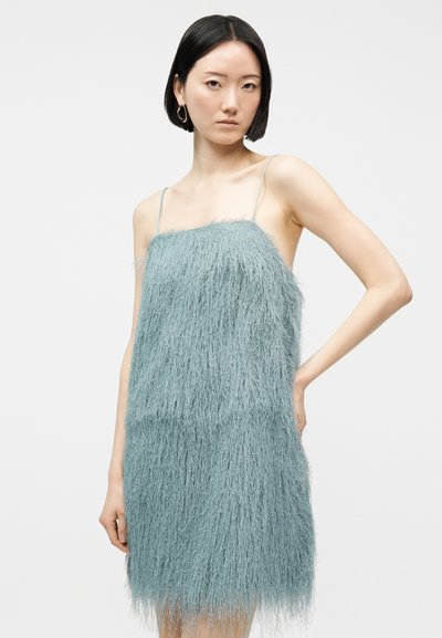 Γυναίκα με κοντά μαύρα μαλλιά, που φοράει ένα ανοιχτό μπλε, χνουδωτό, αμάνικο φόρεμα με λεπτές τιράντες, στέκεται με το ένα χέρι στη μέση.