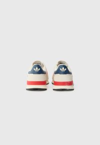Beige sneakers med en marineblå hælpanel, rød accentstribe, sort sål og tre hvide striber som logo på bagsiden.