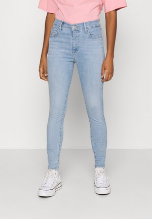 Personne portant un jean skinny bleu clair, des baskets montantes blanches et un t-shirt rose à manches courtes, debout devant un fond uni.