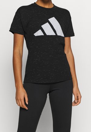 Person iført en sort kortærmet skjorte med et hvidt geometrisk logo på brystet og sorte bukser, stående mod en ensfarvet baggrund.