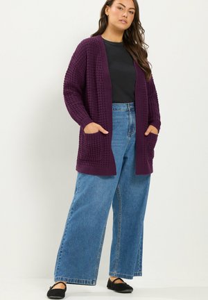 Vrouw draagt een donkerpaarse gebreide cardigan, zwart shirt, wijde blauwe jeans en zwarte platte schoenen, staand met handen in de zakken tegen een lichte achtergrond.
