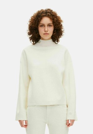 Vrouw met krullend haar, gekleed in een crèmekleurige hoge hals met lange mouwen en bijpassende broek tegen een eenvoudige witte achtergrond.