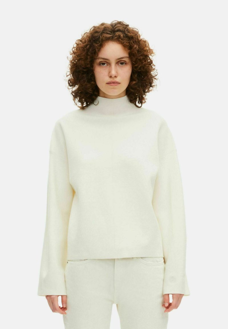 Donna con capelli ricci che indossa un top a maniche lunghe e collo alto color crema e pantaloni abbinati su uno sfondo bianco semplice.
