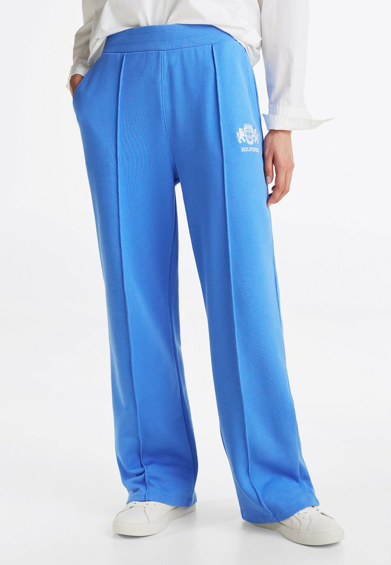 Niebieskie, szerokie spodnie dresowe z ribbowanym paskiem w talii, z delikatnymi fałdami z przodu i logo nadrukowanym na lewej biodrze. Noszone z białymi trampkami.