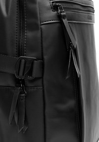 Mochila negra hecha de nailon texturizado con acentos de cuero suave, que cuenta con múltiples cremalleras, correas y hebillas ajustables para un cierre seguro.