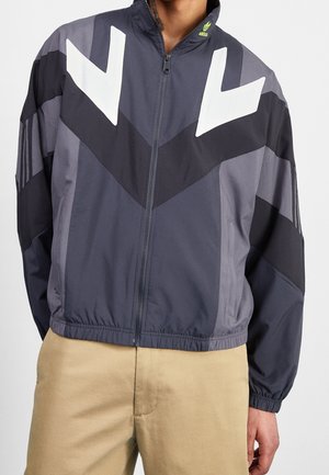 Homme portant une veste zippée gris foncé et noire avec des motifs de flèches blanches sur la poitrine et un pantalon beige, se tenant devant un fond neutre.