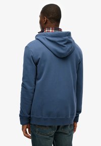 Felpa blu con cappuccio e finitura testurizzata, caratterizzata da una tasca a marsupio e polsini a coste. Indossata sopra una camicia a quadri, con vestibilità comoda.