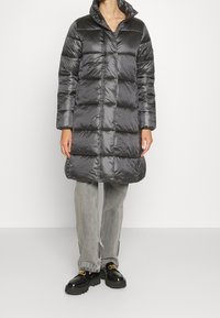 Prošívaný tmavě šedý kabát s výstřihem do V a dlouhými rukávy, s prošitým vzorem. Nošený přes světle šedé džíny a lesklé černé boty.