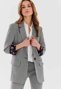 Čierno-biely károvaný blejzer s ružovým akcentom na klopu a farebnými vzorovanými manžetami. V kombinácii s bielou košeľou a zladenými nohavicami.