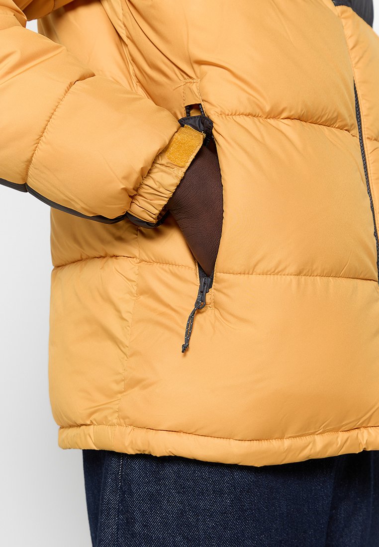 Chaqueta acolchada amarilla con cuello alto, bolsillos con cremallera y detalles en negro. El material tiene una textura suave y un ajuste cálido.