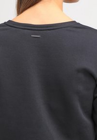 Mörkgrå sweatshirt med rundad halsringning, ribbad textur och en liten varumärkesskylt nära nacken. Slät tyg.