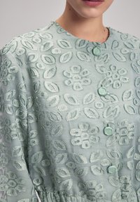 Ljusgrön broderad blus med blommönster, rundade manschetter och dekorativa knappar. Genomskinlig tyg med en texturerad yta.