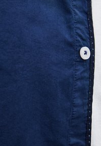 Materiał koszuli w kolorze granatowym o gładkiej fakturze, z białym plastikowym guzikiem i kontrastującym szwem wzdłuż szwu.