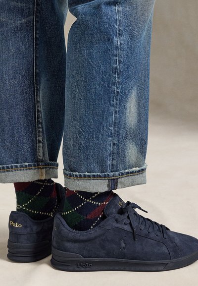 Ben som bär uppvikta blå jeans, mönstrade mörka strumpor och marinblå Polo Ralph Lauren-sneakers med snören på en ljus yta.