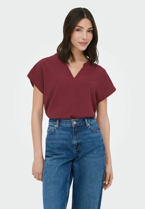 Vrouw met donker haar die een korte mouw burgundy V-hals top en een hooggetailleerde blauwe spijkerbroek draagt, staand tegen een effen achtergrond.