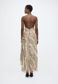 Γυναίκα με κοντά μαλλιά που φοράει ένα μακρύ μέχρι τους αστραγάλους φόρεμα με σχέδιο φιδιού και μαύρες πλατφόρμες με τακούνι, όρθια με την πλάτη προς το μέρος μας σε λευκό φόντο.