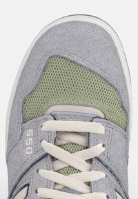 Baskets en daim gris avec panneaux en maille verte, lacets crème, marque visible sur le côté et semelle en caoutchouc blanche.