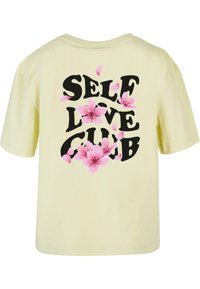 Žluté tričko s krátkým rukávem s nápisem "SELF LOVE CLUB" v černé barvě, doplněné růžovými květinovými vzory. Měkký bavlněný materiál.
