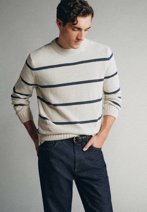 Jeune homme portant un pull blanc à rayures horizontales noires et un jean bleu foncé, debout avec une main dans la poche devant un fond uni.