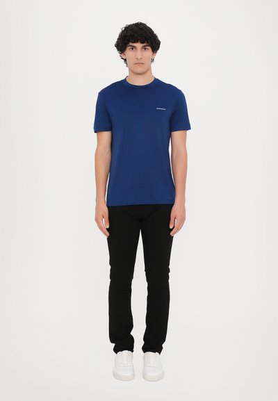 Tamsiai mėlynas apvalios ausies marškinėlis iš lengvos medžiagos, derinamas su juodais, priglundančiais kelnais ir baltais sportiniais batais. Marškinėliai turi nedidelį logotipo detalę.