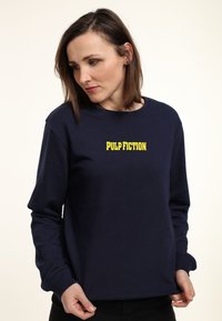 Marineblauwe sweatshirt met lange mouwen, ronde hals en gele "PULP FICTION" tekst op de borst. Gemaakt van een zachte katoenmix.
