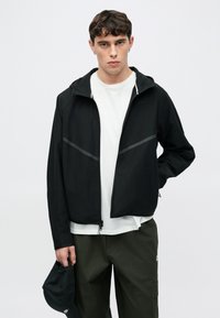 Czarna lekka kurtka z kapturem, z szarymi paskami jako akcentami, pełny zamek błyskawiczny i gładka faktura. Nosi się ją na białą koszulkę.