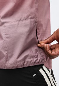 Main tirant la fermeture éclair sur la poche d'une veste mauve au-dessus d'un pantalon noir avec des rayures blanches.