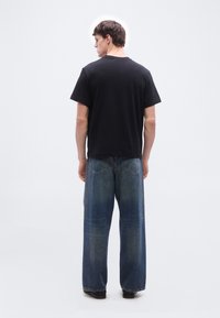 Άνδρας που στέκεται γυρισμένος προς τα πίσω, φορώντας μαύρο κοντομάνικο μπλουζάκι και φαρδιά μπλε τζιν, μπροστά σε απλό λευκό φόντο.