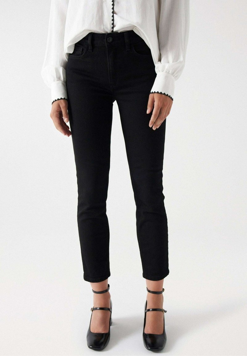 Jean noir slim en denim, taille haute, cinq poches, avec un léger éclat, associé à des talons noirs et une blouse blanche.