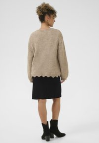 Beige gestrickter Pullover mit gewelltem Saum, über einem schwarzen Rock getragen. Das Outfit wird mit schwarzen, hochhackigen Stiefeletten abgerundet.
